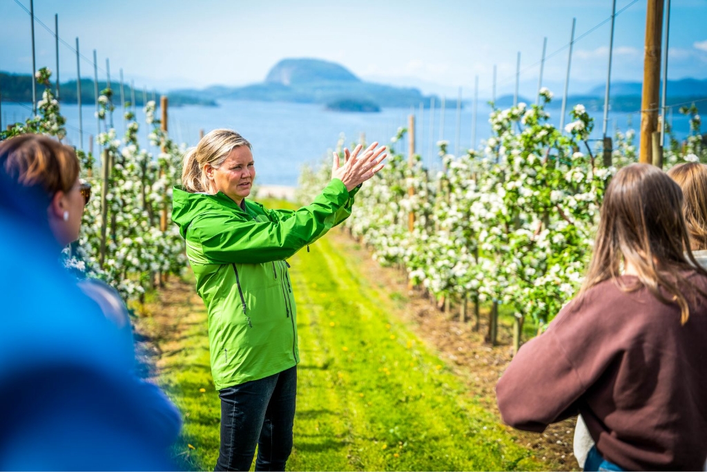 Ei kvinne står framfor mange frukttre og snakkar til ei gruppe mennesker. Foto.