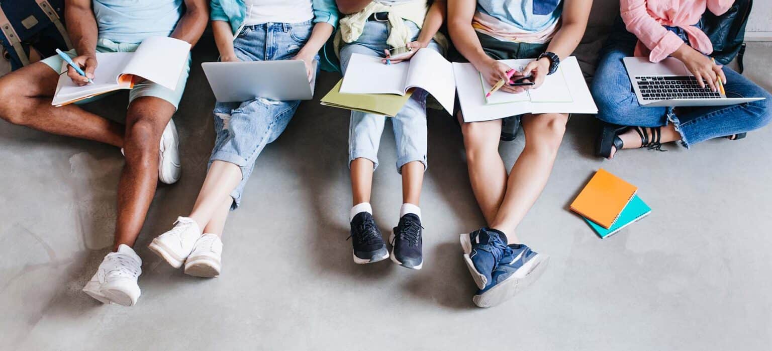 Fem ungdommar sit på golvet med PC og notatbøker i fanget. Foto.