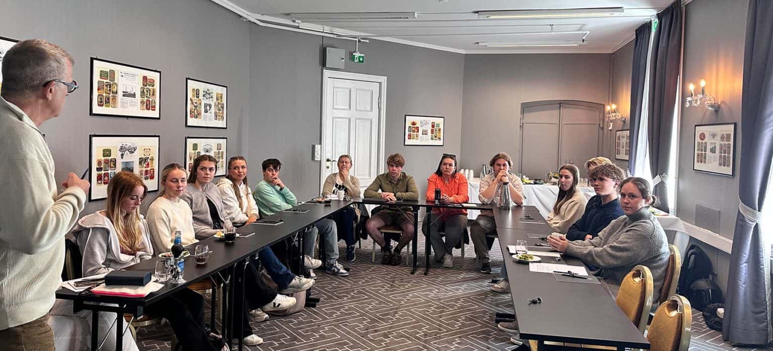 13 ungdommar sit rundt eit bord i hestesko og følger med på ein som underviser. Foto.