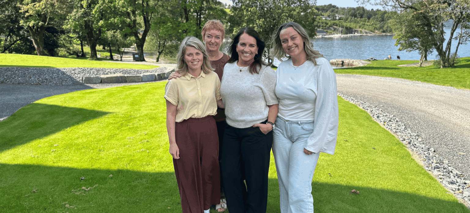 Fire kvinner står samla ute på graset og smiler til kamera.